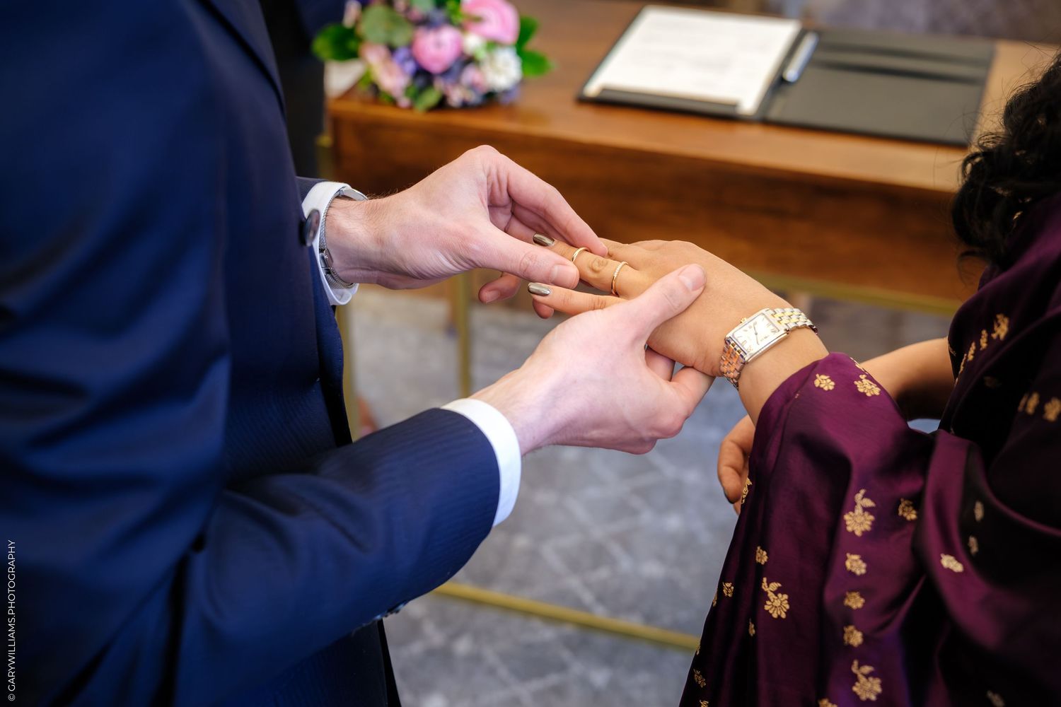 Close up of hands exchanging wedding rings during a traditional ceremony with a burgundy accent color.