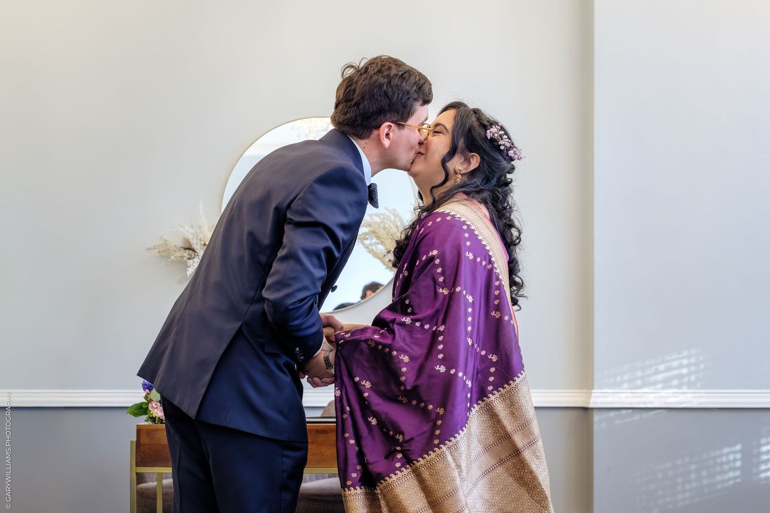 A couple shares a romantic kiss at their wedding ceremony, with the bride wearing a purple dress and groom in dark suit.
