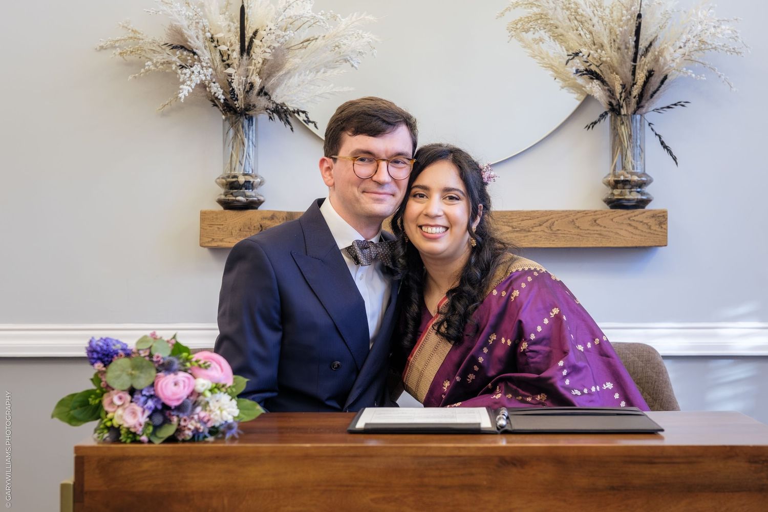 A couple poses together at a wedding venue with decorative palm fronds and floral arrangements in the background.