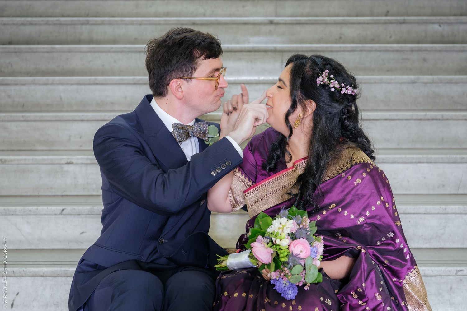 A couple in formal attire share a tender moment on marble steps, with the bride holding a floral bouquet.