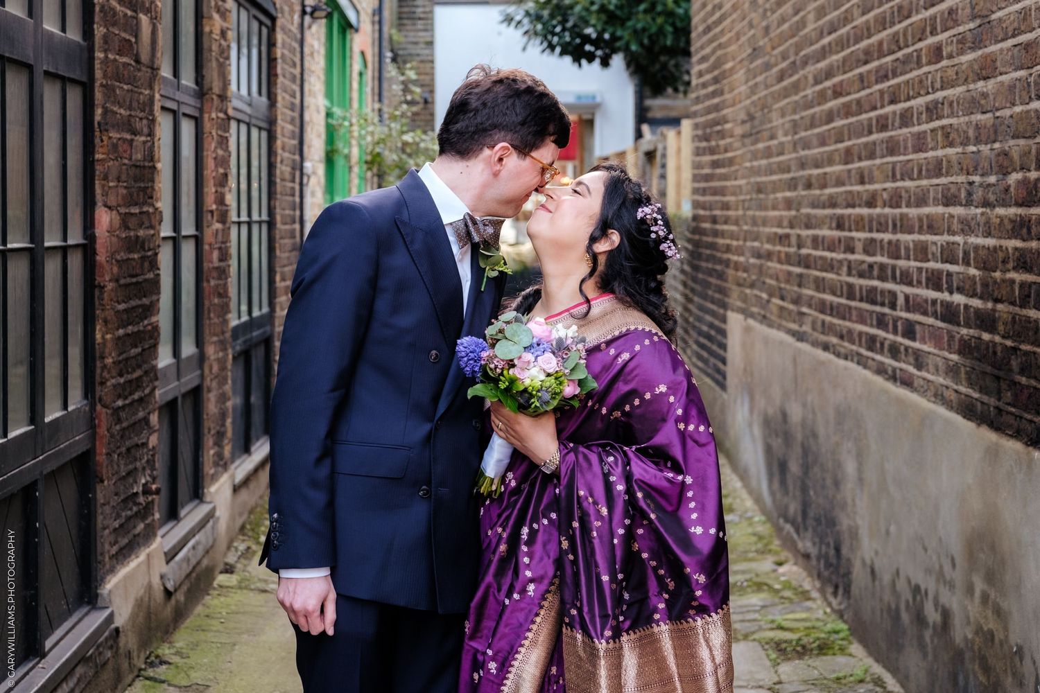 A couple shares a romantic moment in an alley wearing formal attire with a purple sequined dress and navy suit.