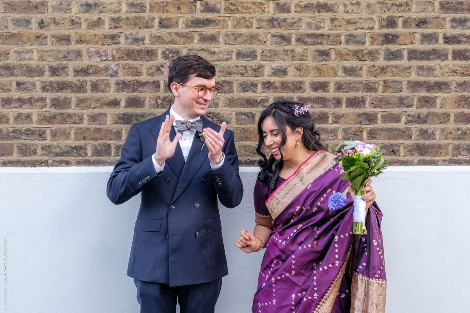 A couple shares a lighthearted moment together against a brick wall, with one wearing a navy suit and one in a purple sari.