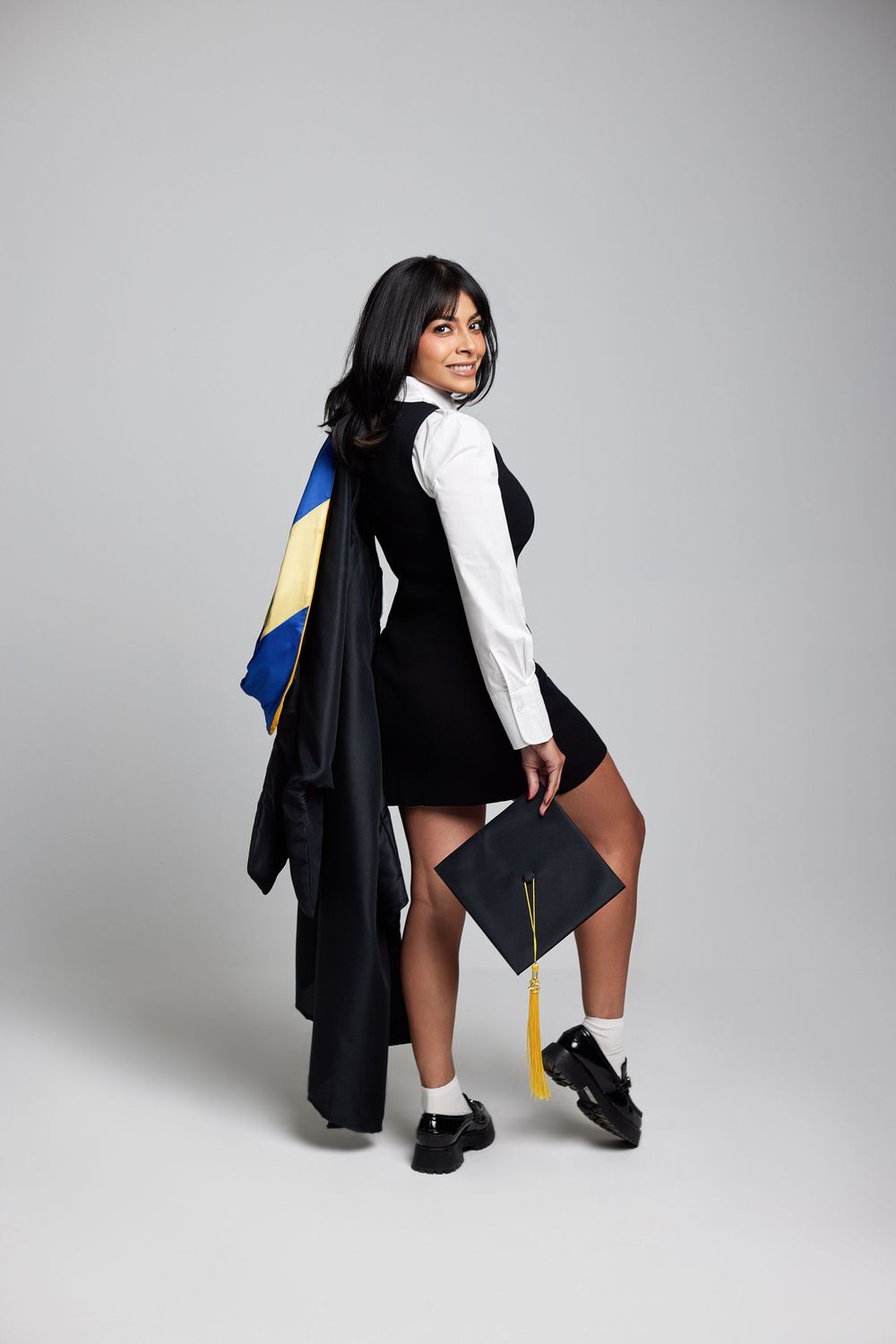 Graduate poses playfully in cap and gown against light grey studio background.