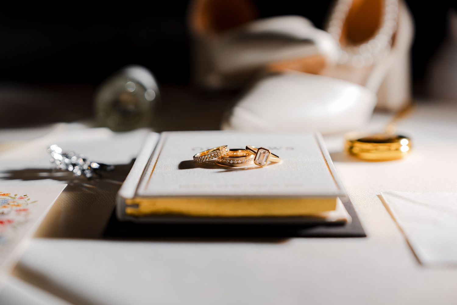 Gold wedding rings resting on a small book with wedding shoes and accessories in the background.
