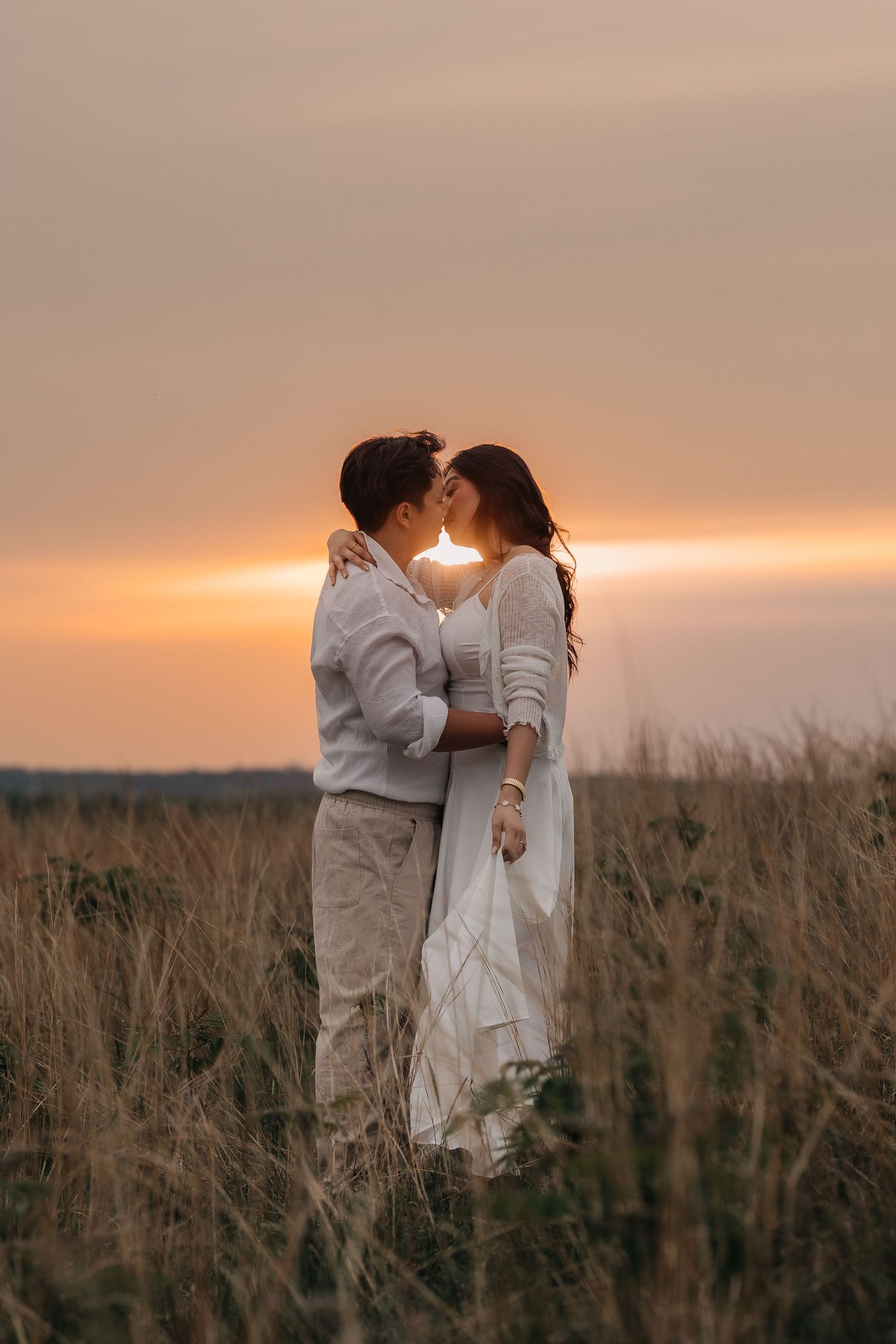 A couple in white outfits share an intimate moment at sunset in a rustic field with tall grass.