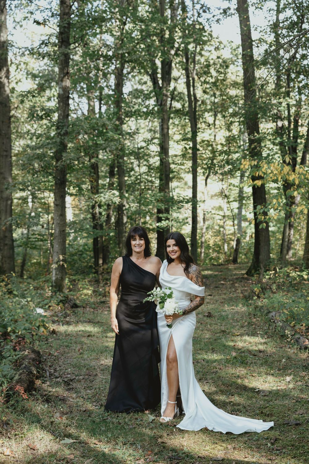 Wedding party poses in a sunlit forest setting with bridesmaids in dark dresses and bride in white gown.
