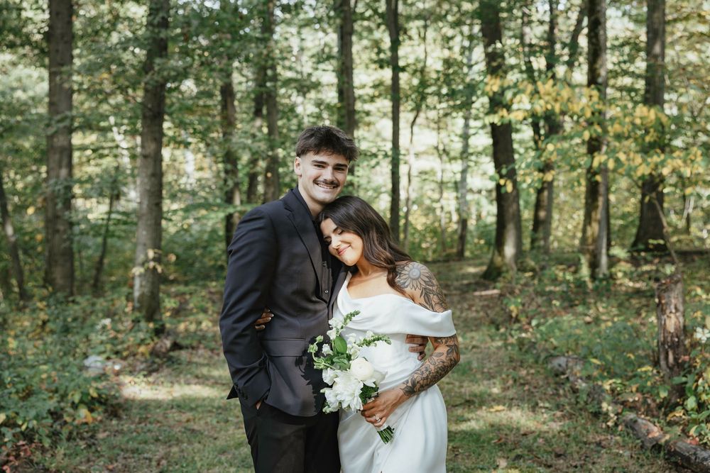 A romantic wedding couple embraces in a sunlit forest setting while holding a white floral bouquet.