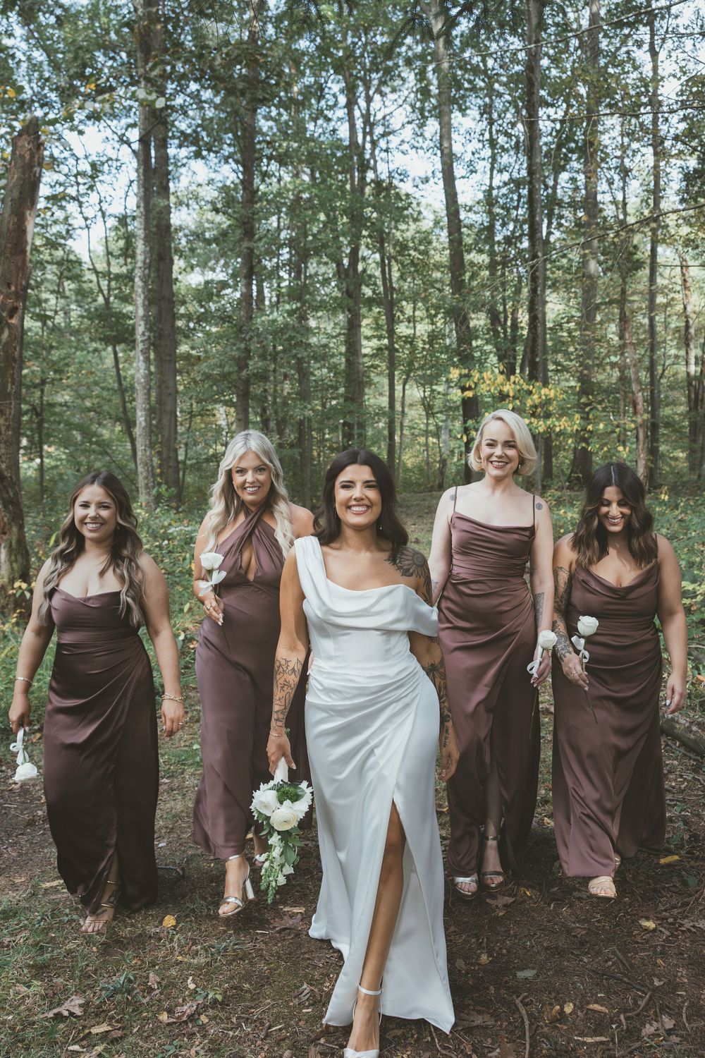 A bridal party poses in a forest setting wearing brown bridesmaid dresses and a white wedding gown.