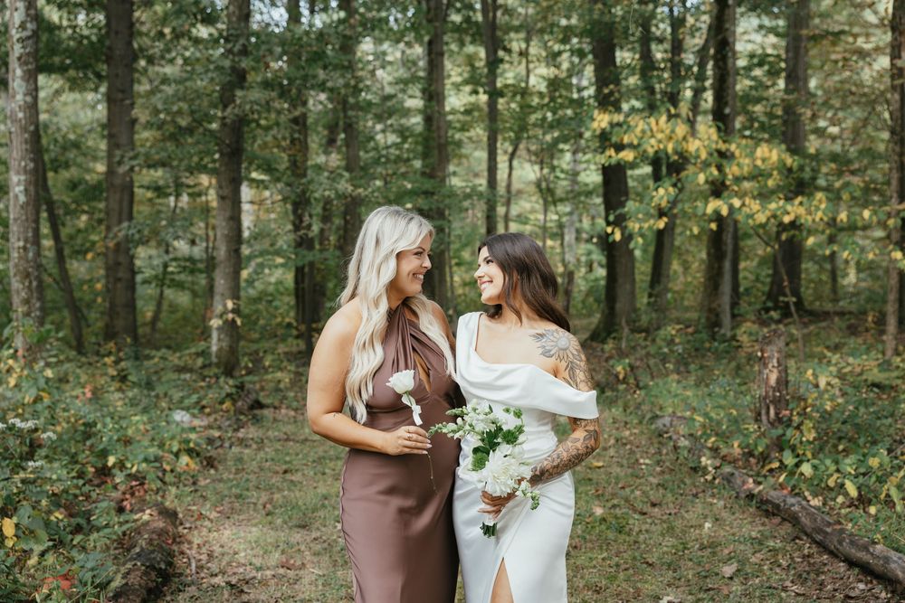 Two people in white dresses share an intimate moment in a sunlit forest setting.