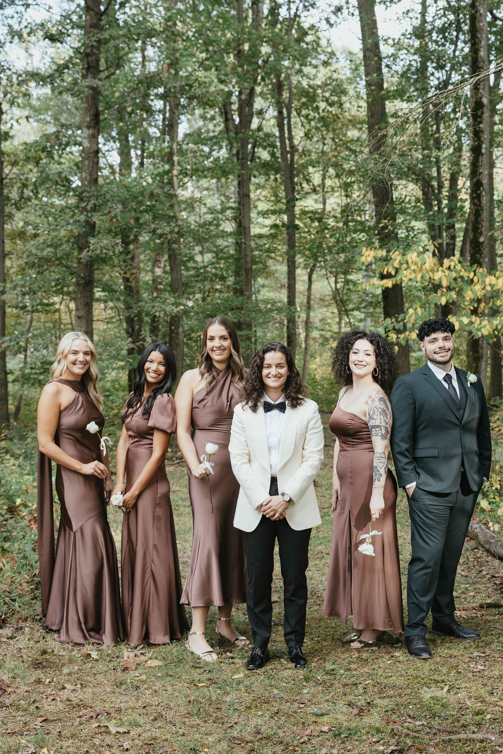 Wedding party members stand together in a wooded area wearing brown and dark formal attire.