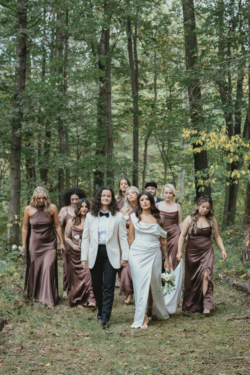 Wedding party in brown bridesmaid dresses and white gown walking together through a wooded forest setting.