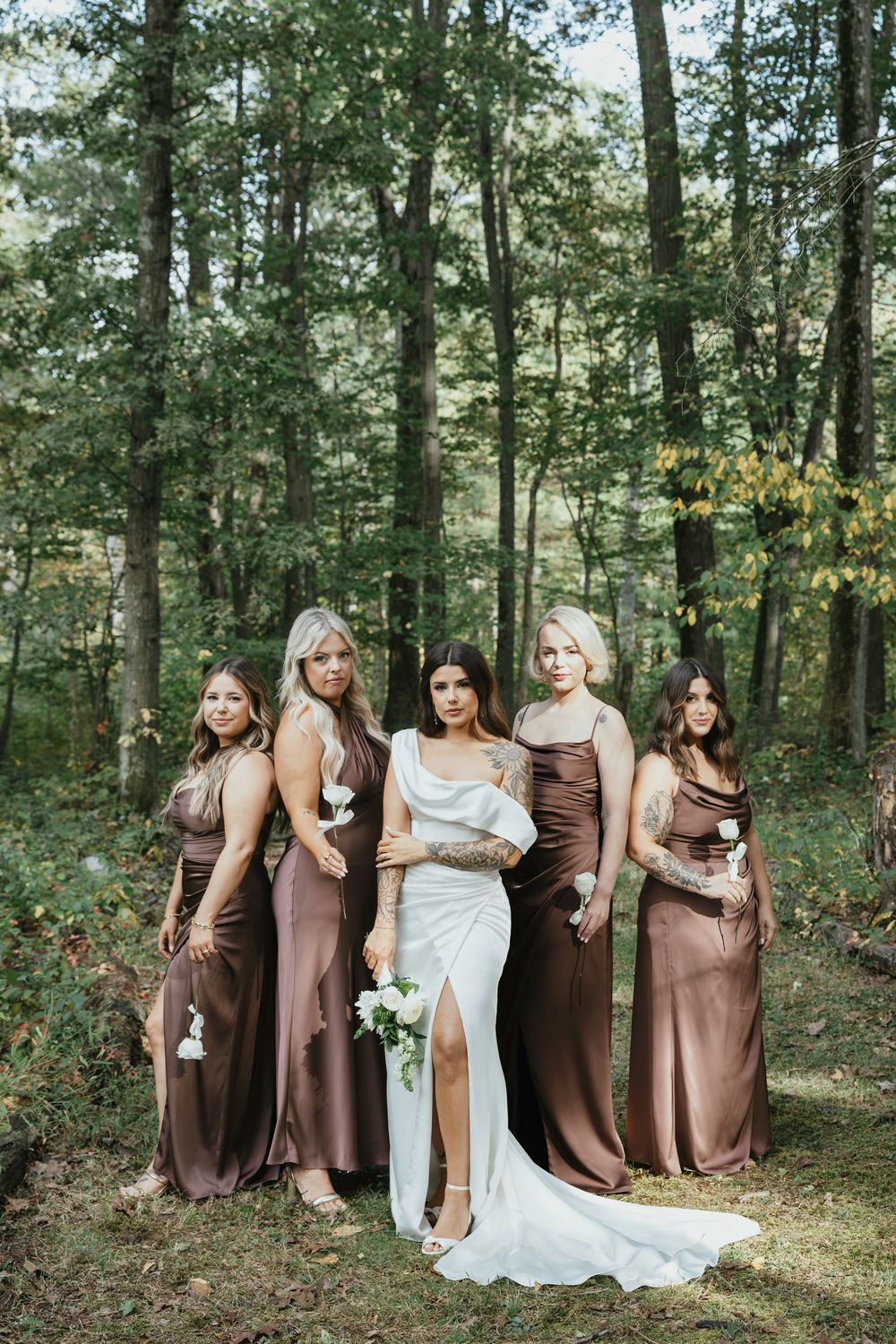 Wedding party poses playfully in a line among trees wearing chocolate brown dresses and white bridal gown.