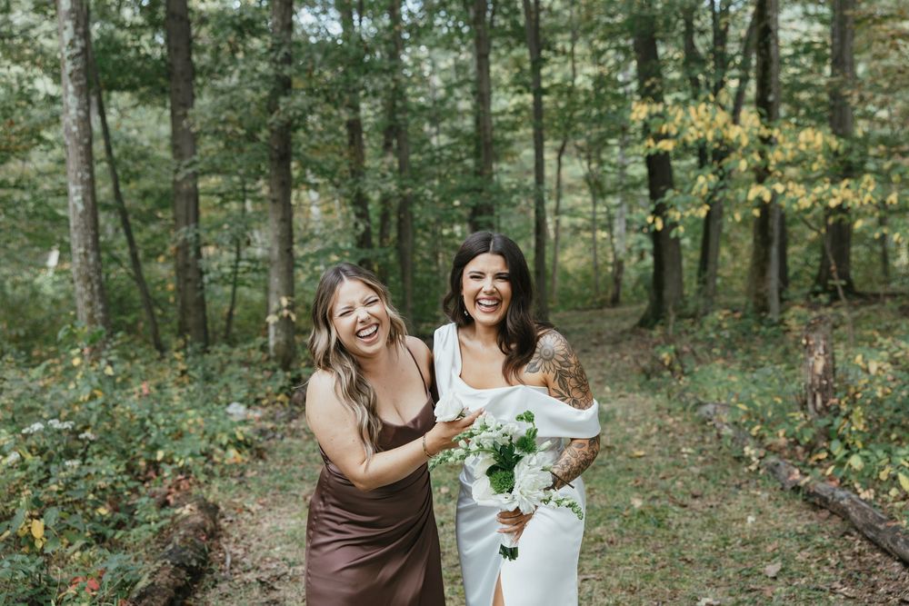 Wedding party members share candid laughing moments in a forest setting wearing brown and white dresses.