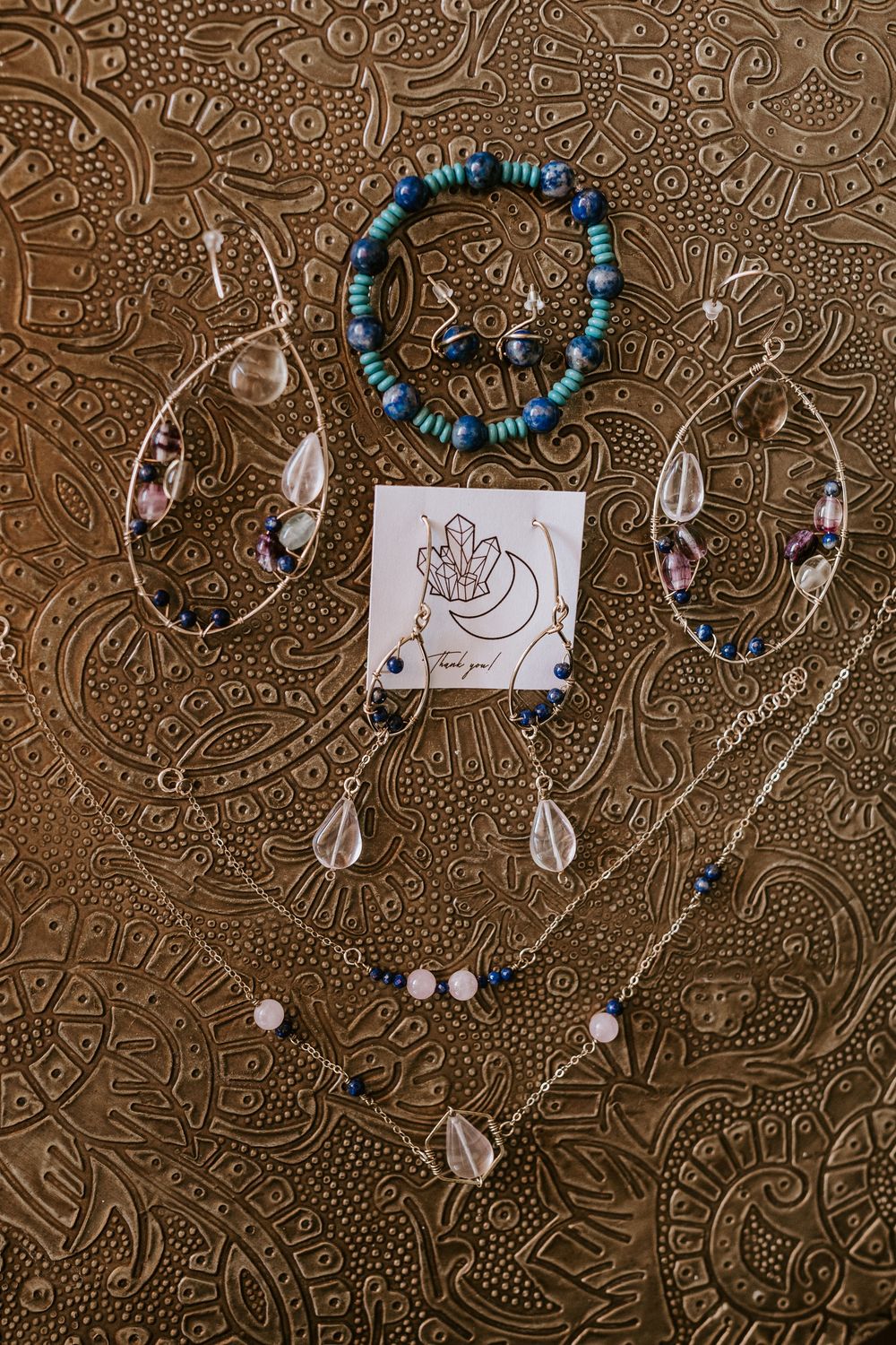 Bridal jewelry flat lay featuring crystal earrings, blue beaded bracelet, and delicate necklaces on gold surface.