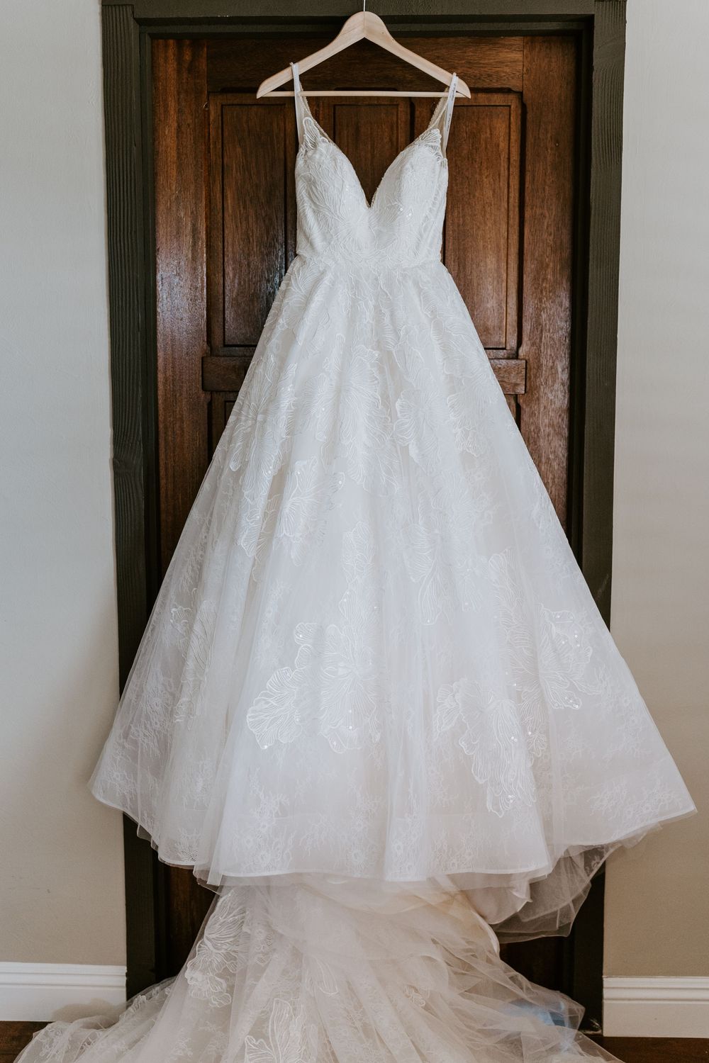 Elegant white lace ball gown wedding dress with deep V-neckline and long train hanging on wooden door.