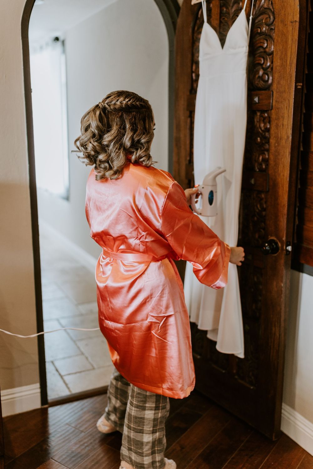 Young flower girl in coral satin robe steaming a white wedding dress hanging on an ornate wooden door.