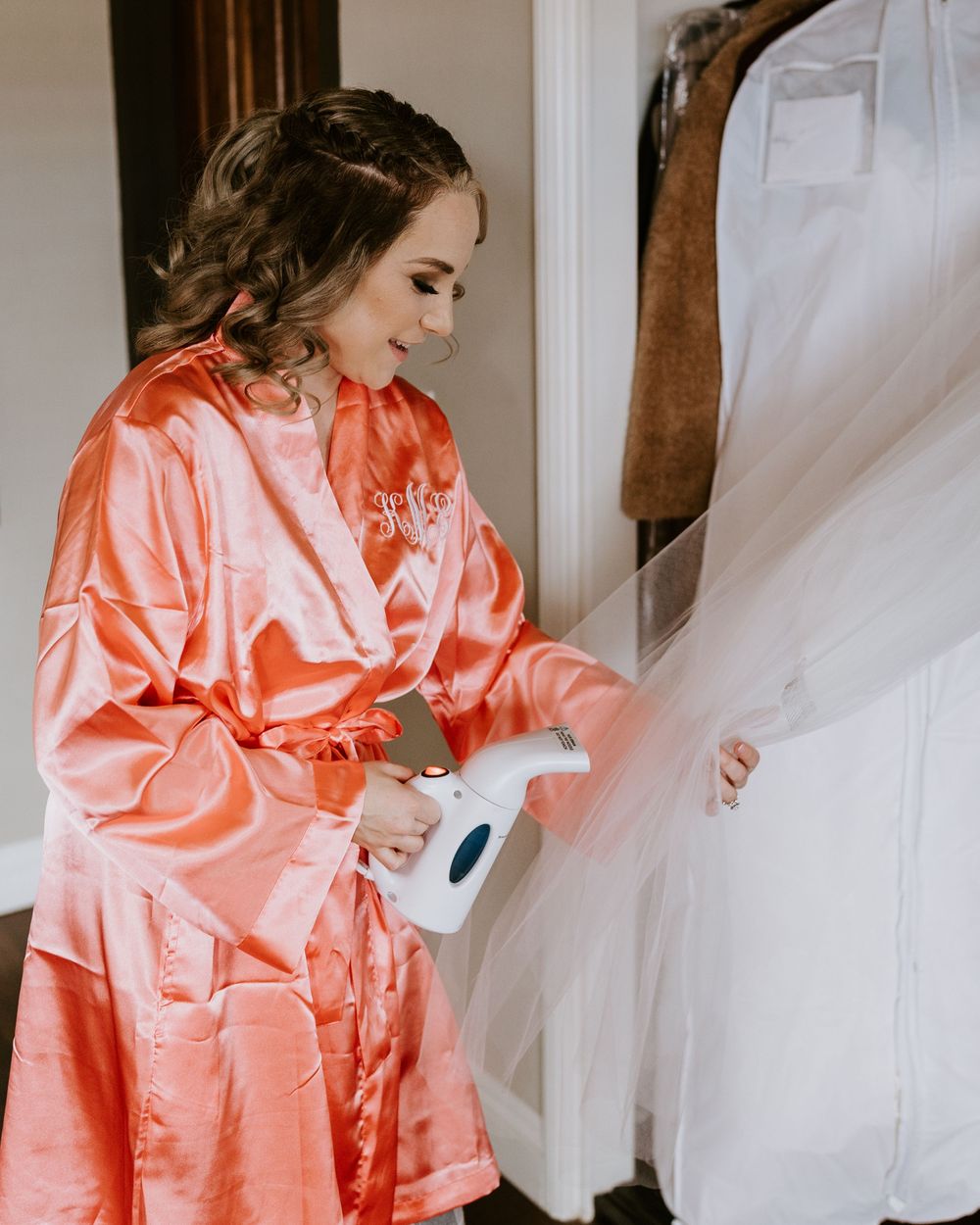Bridesmaid in coral satin robe steaming white wedding dress on hanger.