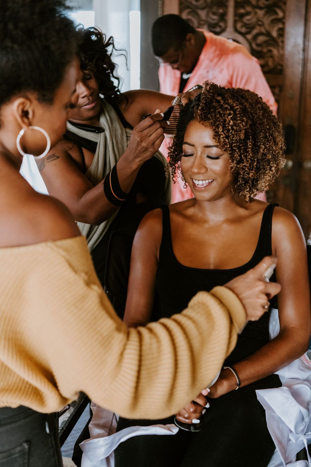 Bride smiling joyfully while stylists do her hair and makeup during wedding preparations.