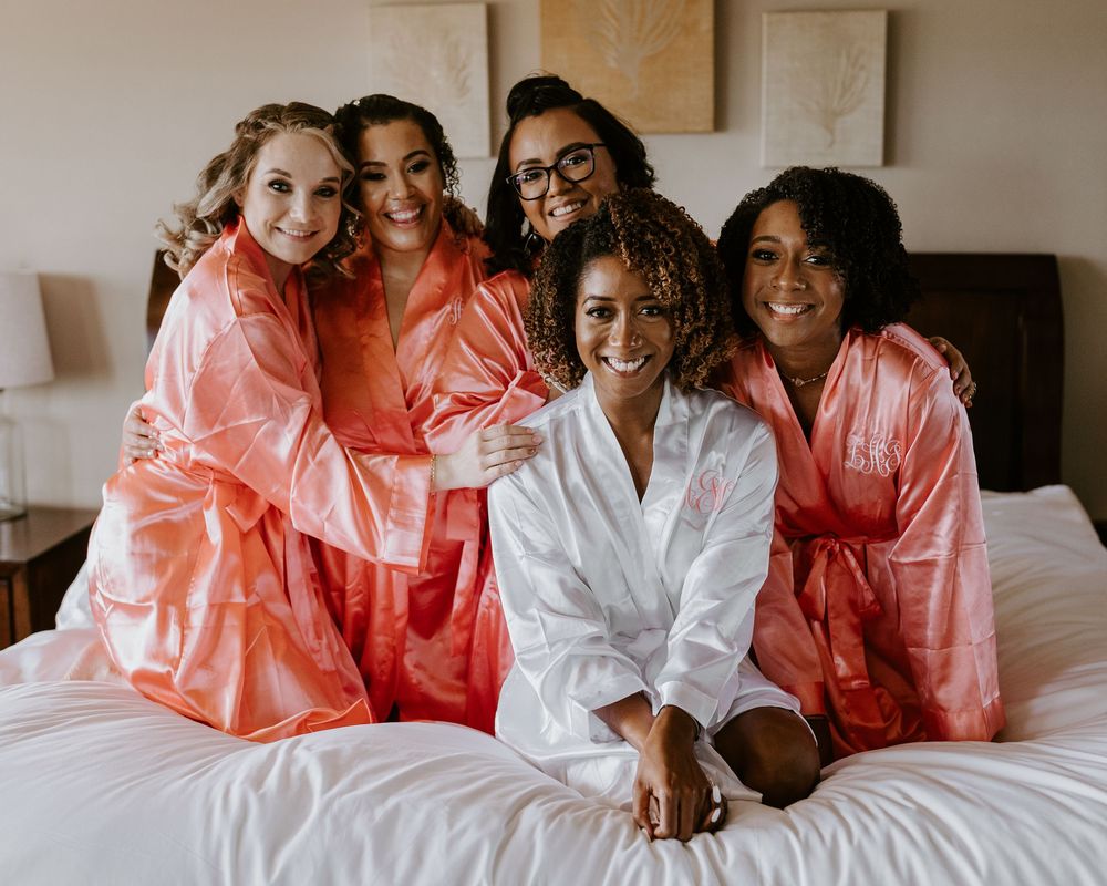 Bride in white satin robe posing with four bridesmaids in coral robes on white hotel bed.