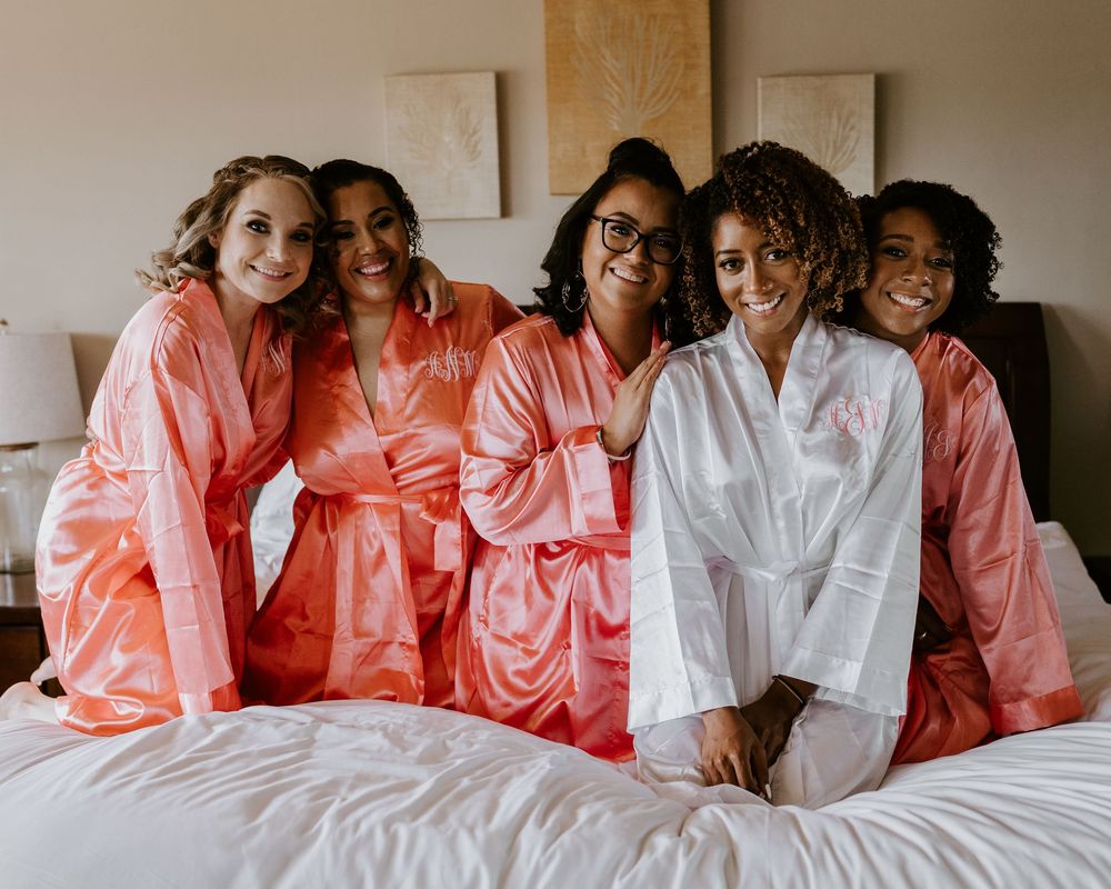 Five bridesmaids in coral and white satin robes sitting together smiling on hotel bed.