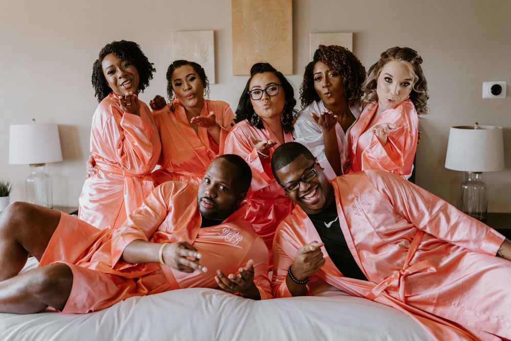 Bride, bridesmaids, and groomsmen in satin robes laughing together on white hotel bed.