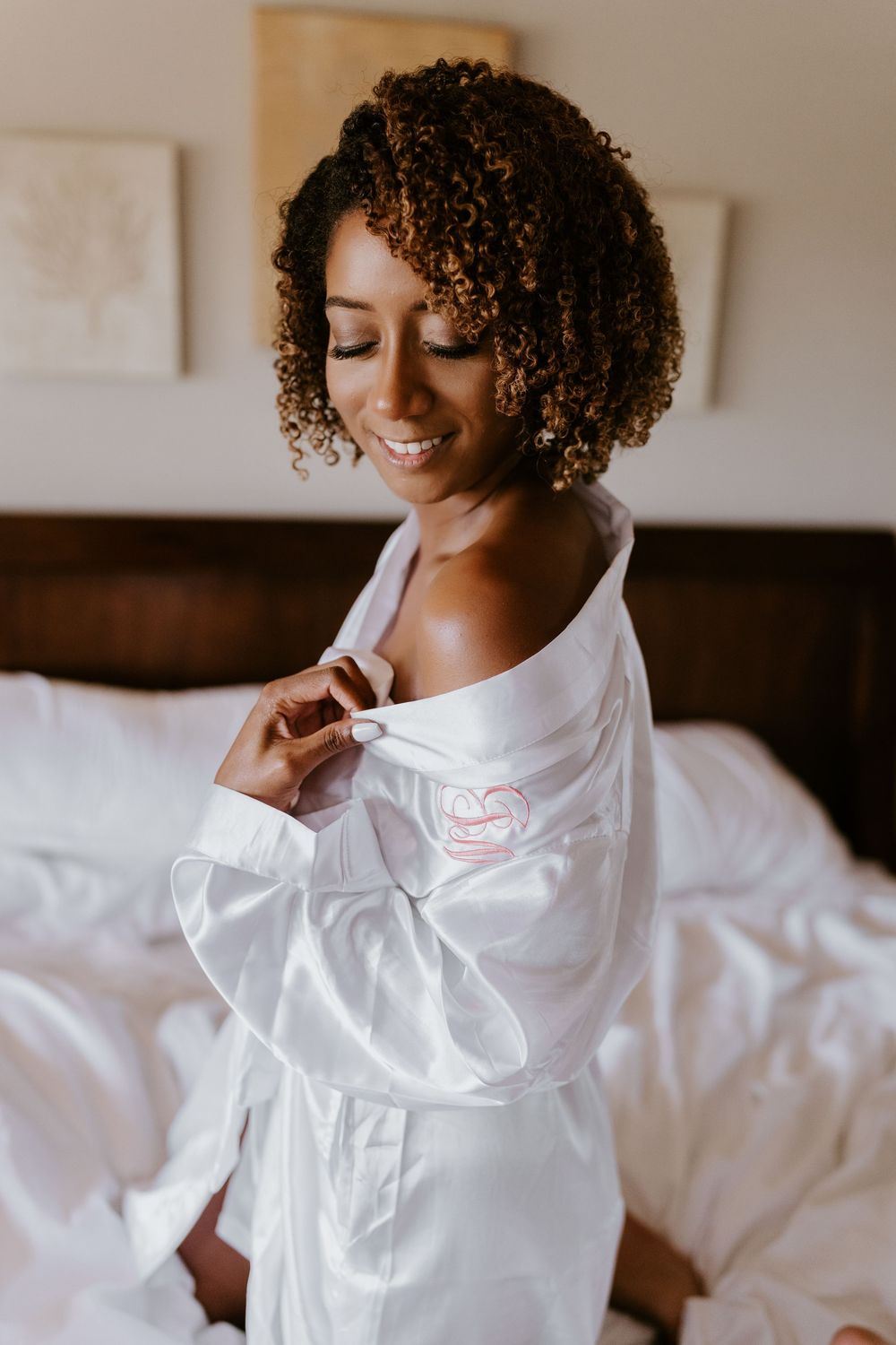 Smiling Black bride in white satin monogrammed robe sitting on bed with natural curly hair.