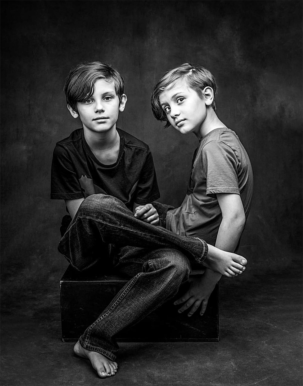 Black and white artistic photo series showing two children sitting together in casual poses against a dark background.