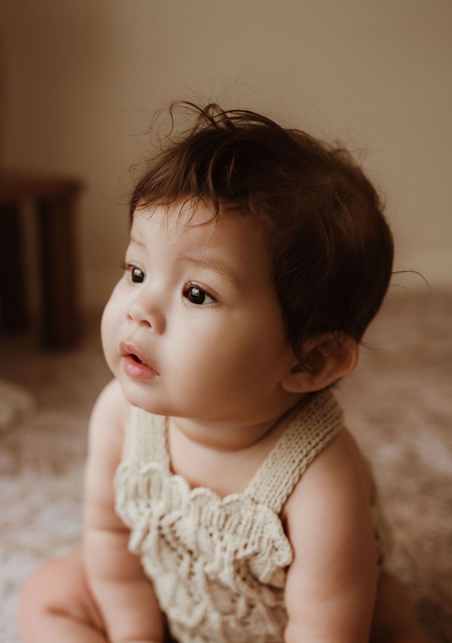 Baby in white outfit sitting on textured cream rug against minimalist white wall backdrop.