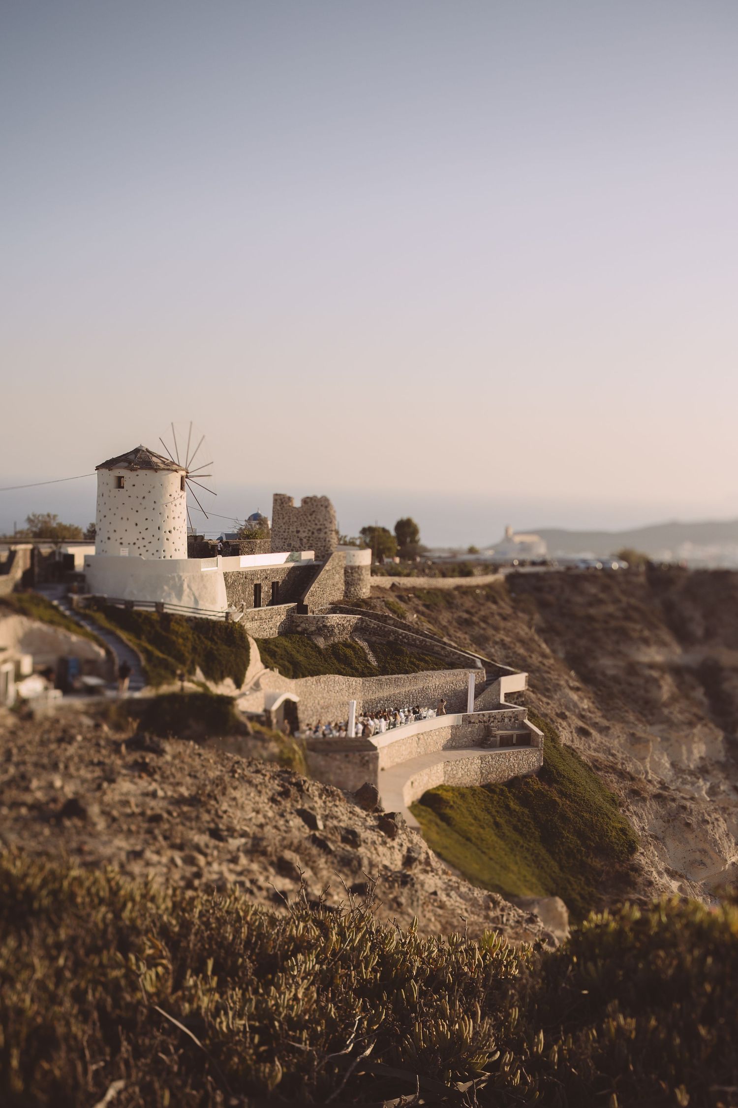 White El Viento windmills and buildings perched on rocky cliffs overlooking the sea at sunset in Santorini, Greece.