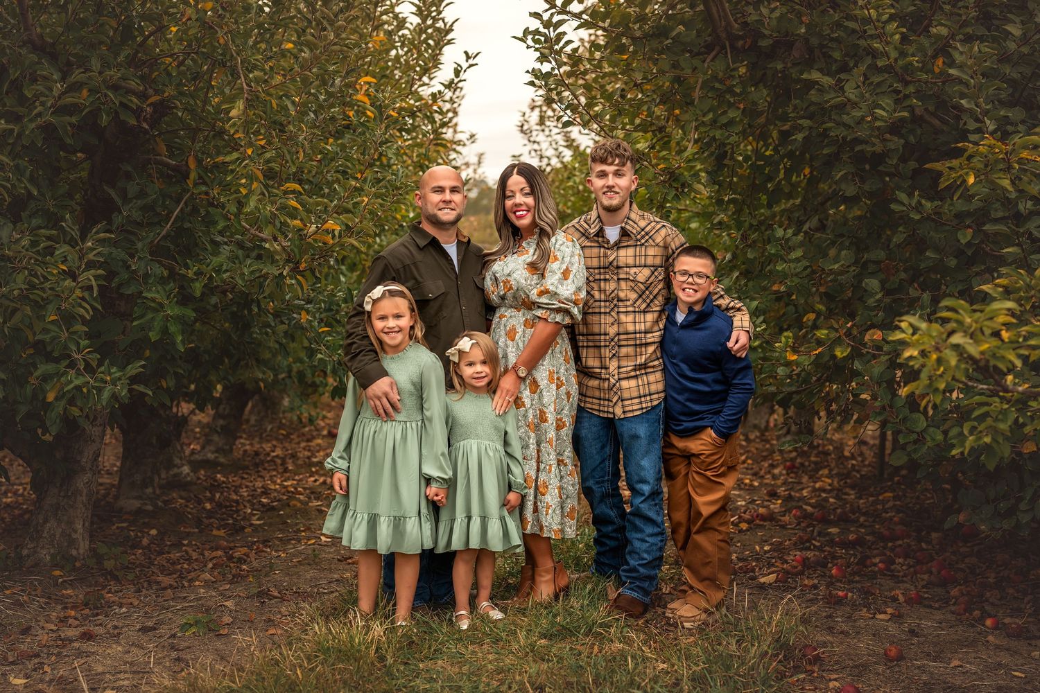 A group stands together in a rustic orchard setting during autumn with matching sage green dresses and casual attire.