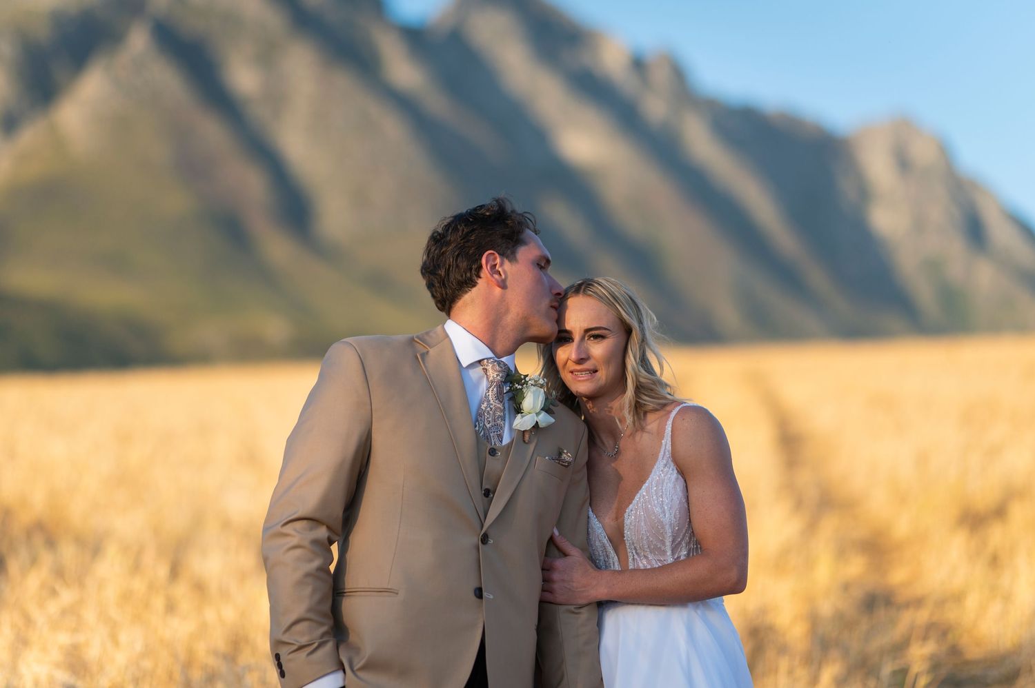 Bride and groom share a romantic moment in a golden field with dramatic mountain backdrop on their wedding day.