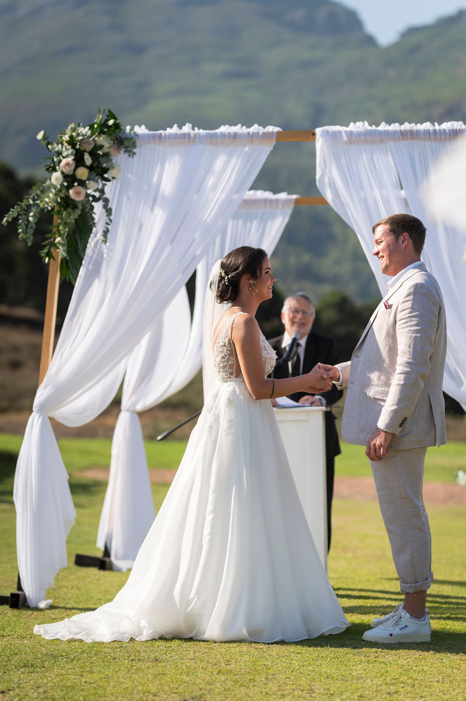 Bride and groom exchange vows under a white draped arch with floral decor on a green lawn with mountains behind.