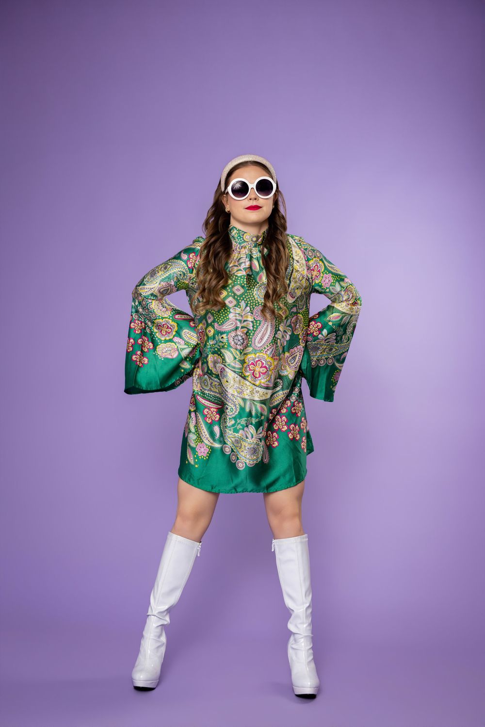 Retro-styled fashion shoot with white sunglasses, green floral dress and boots against purple studio background.