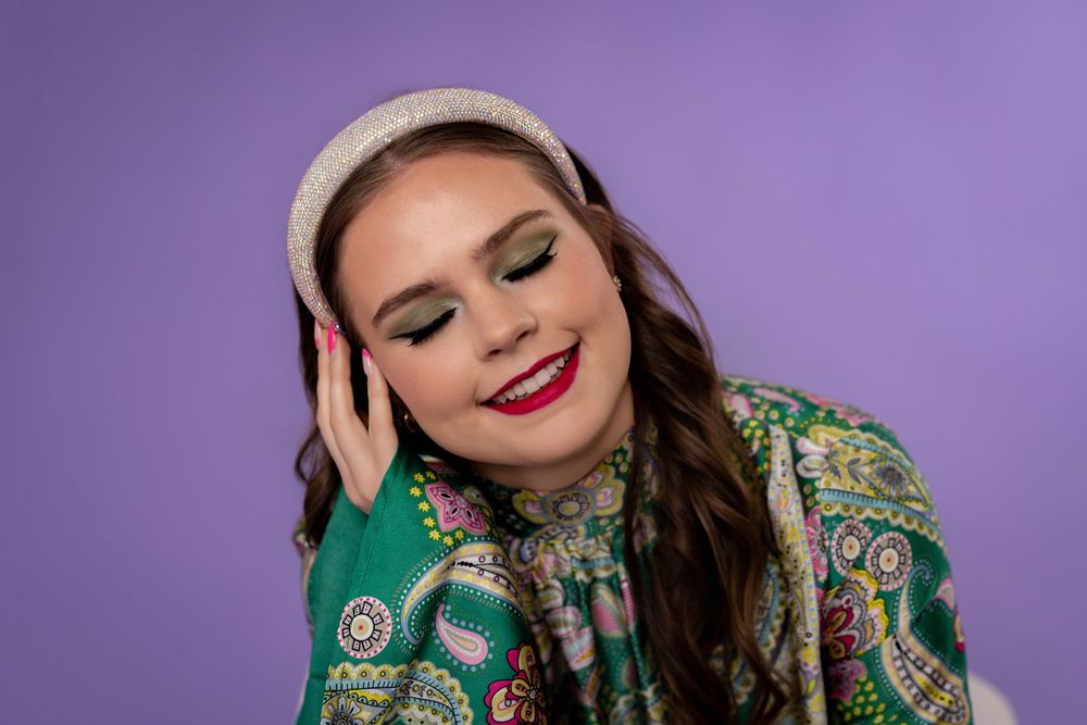 A person wearing a green sequined jacket and dramatic makeup poses against a purple background in a fashion photoshoot.