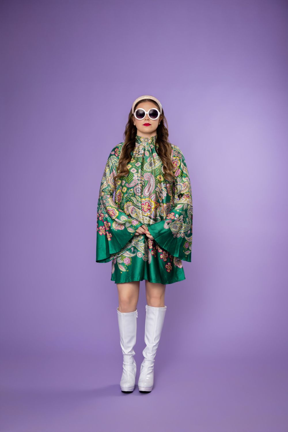 Fashion model in green floral dress with white boots and sunglasses poses against purple background.