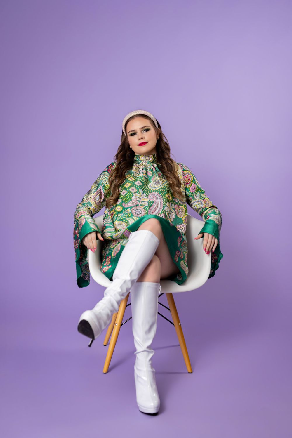 Stylish figure lounges in white chair wearing emerald patterned robe and tall boots against lavender backdrop.