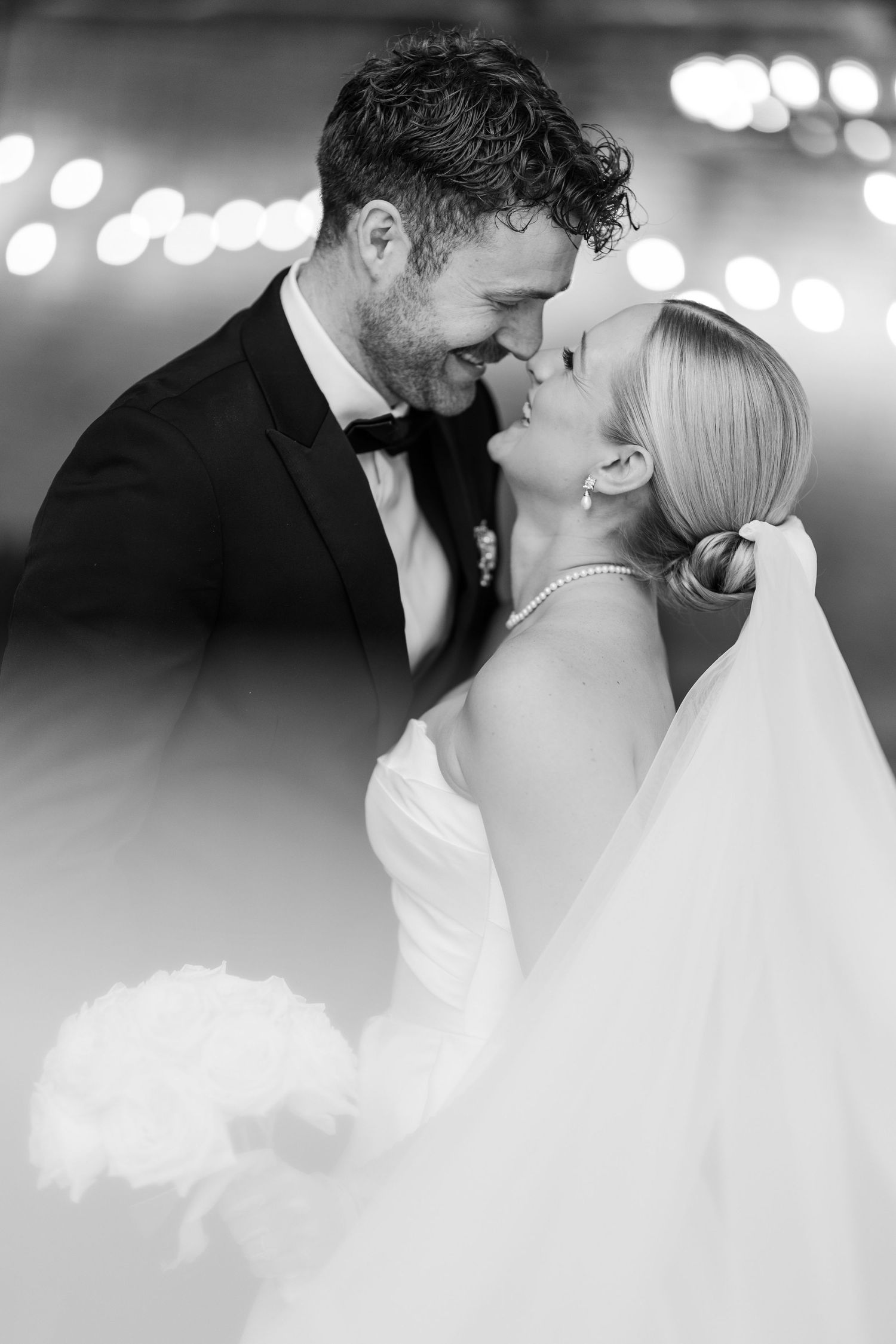 A romantic wedding couple shares intimate moments in a warmly lit venue with string lights glowing in the background.
