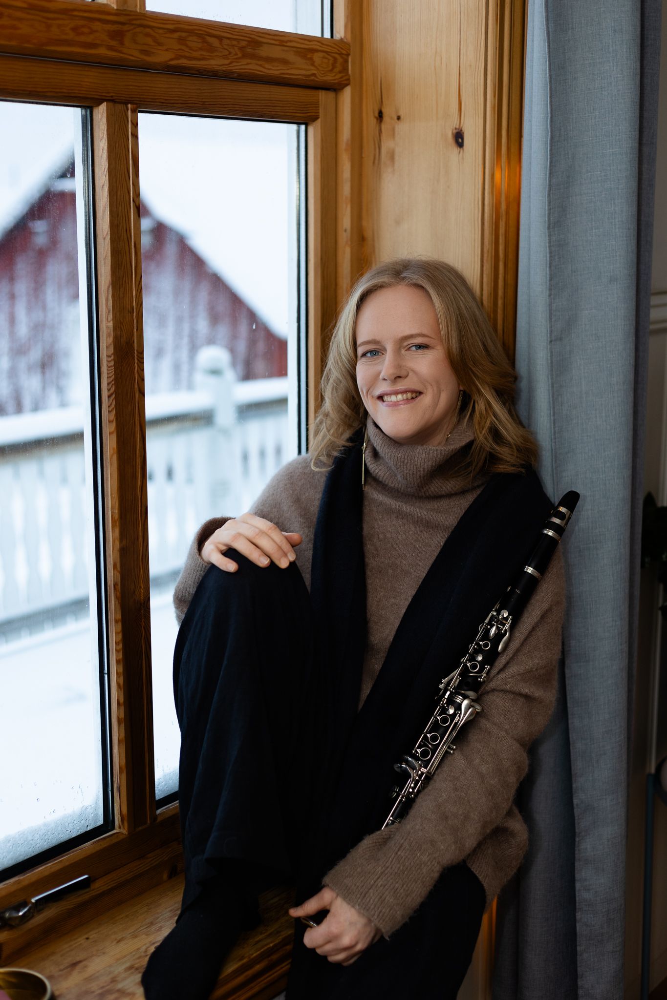 Musician in brown turtleneck sweater sits by window holding clarinet with red barn visible in snowy background.