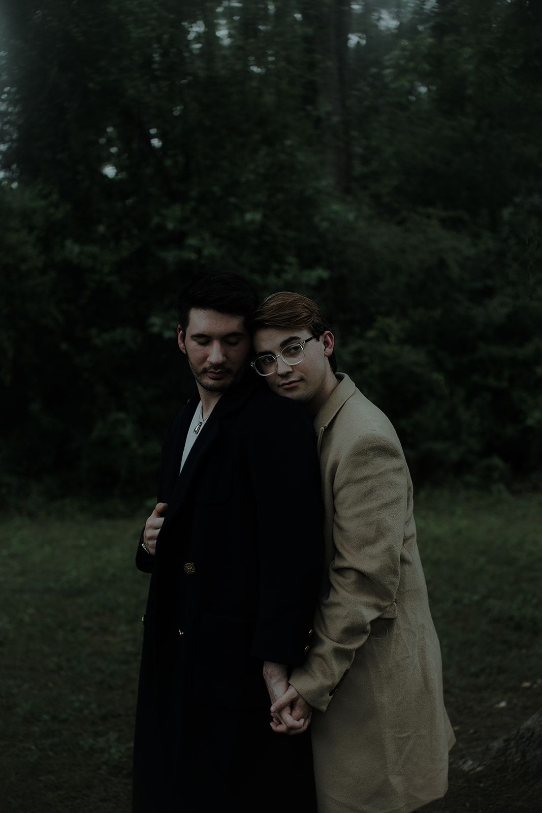 Two people in formal attire share a tender moment outdoors against a dark forest backdrop.