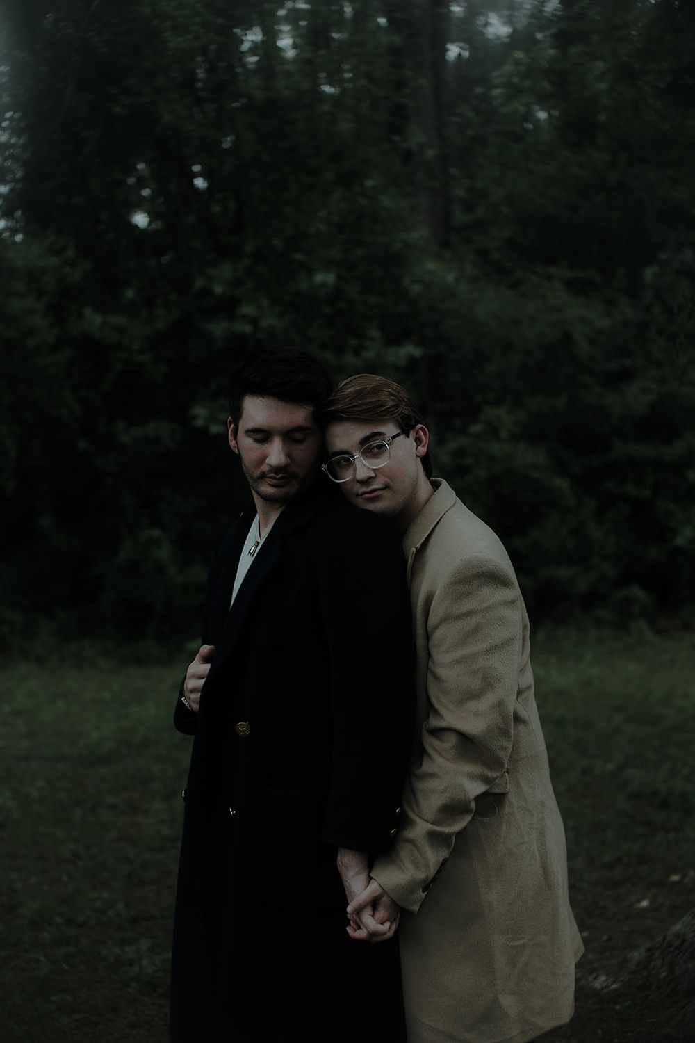 Two people in formal attire share a tender moment outdoors against a dark forest backdrop.