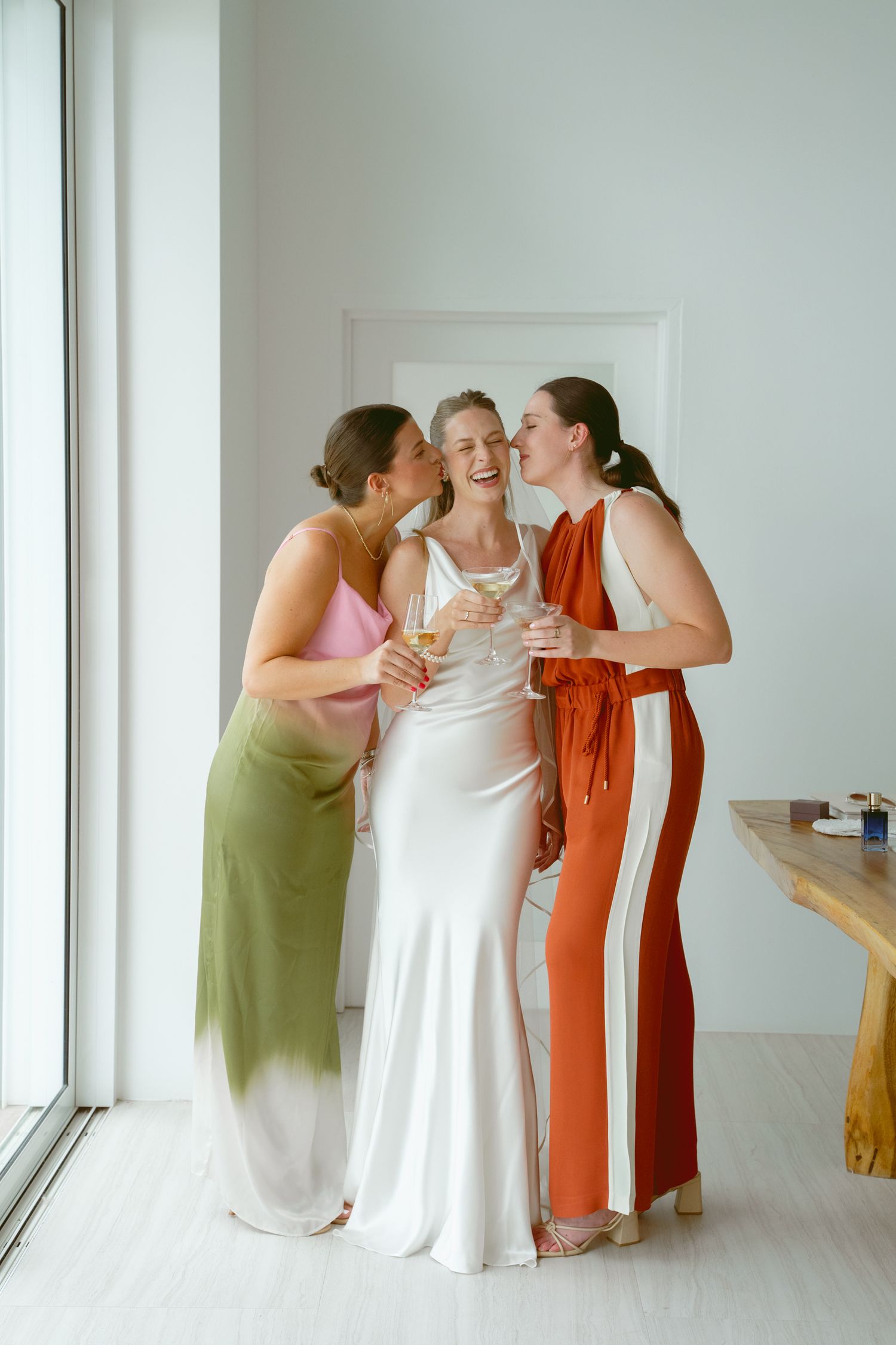 Three people in colorful dresses share an affectionate moment in a bright white room.