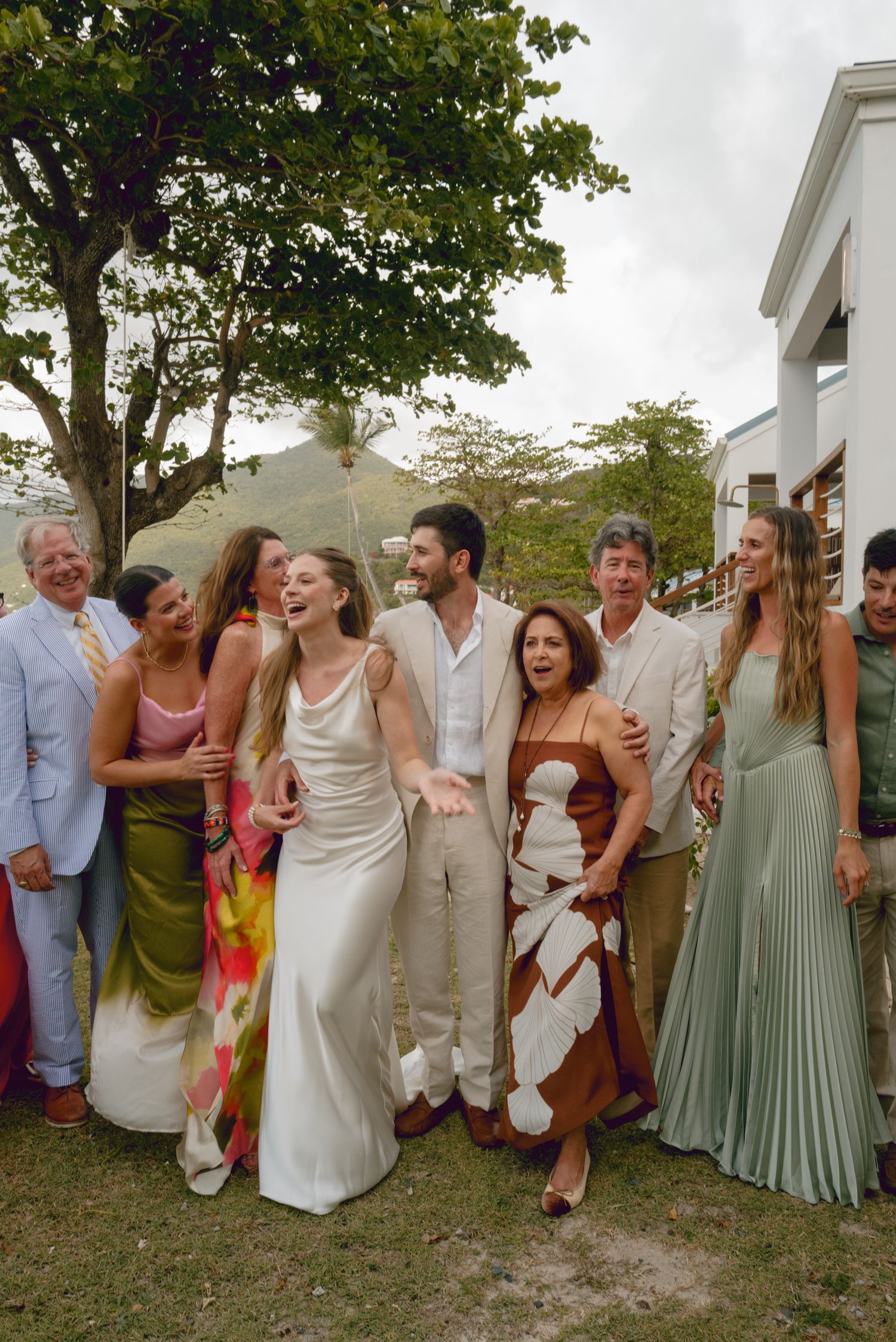 Wedding party laughing together outdoors with guests in formal attire at a summer celebration.