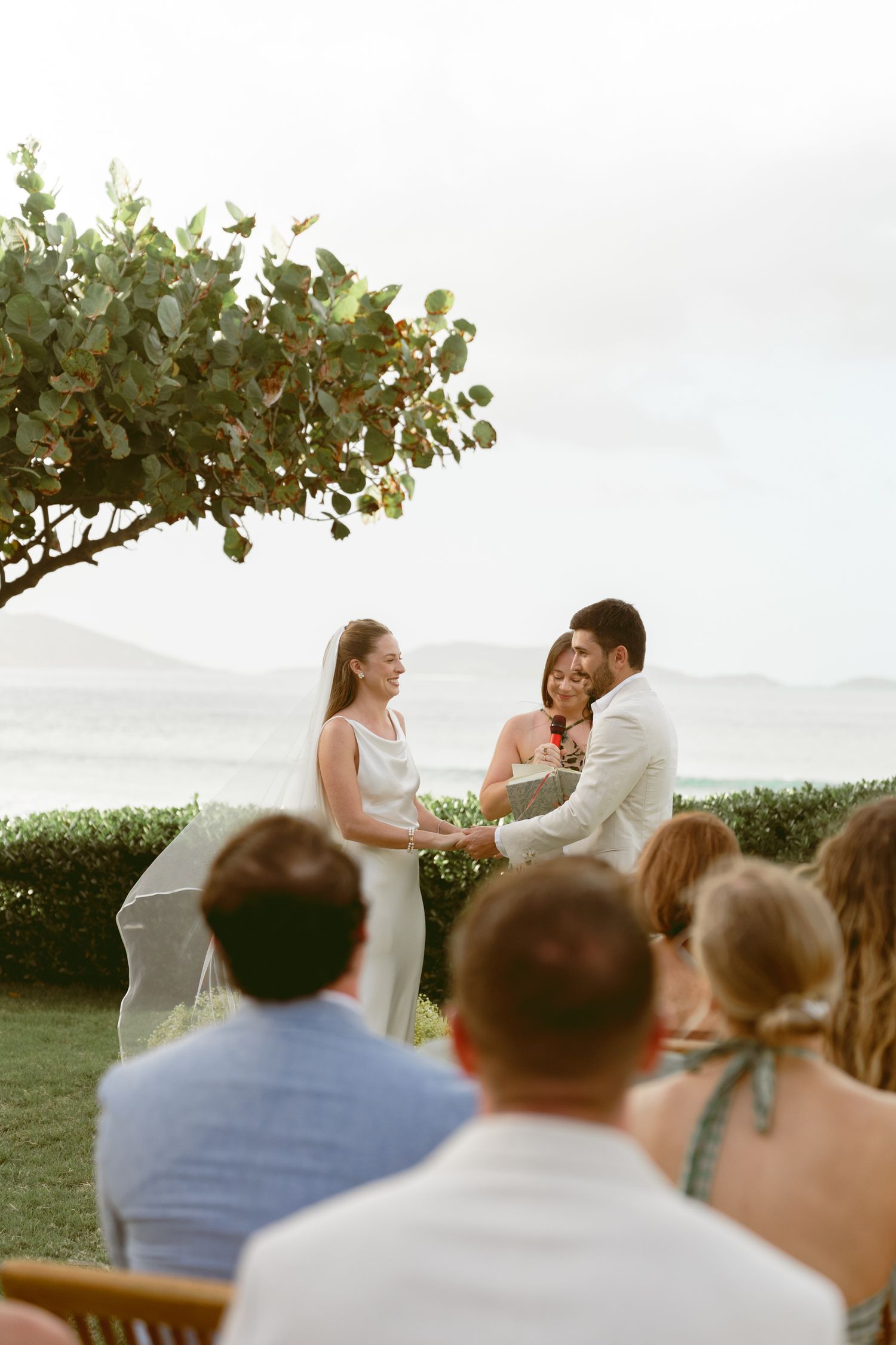 Wedding ceremony guests seated outdoors watching a couple exchange vows under tropical foliage.
