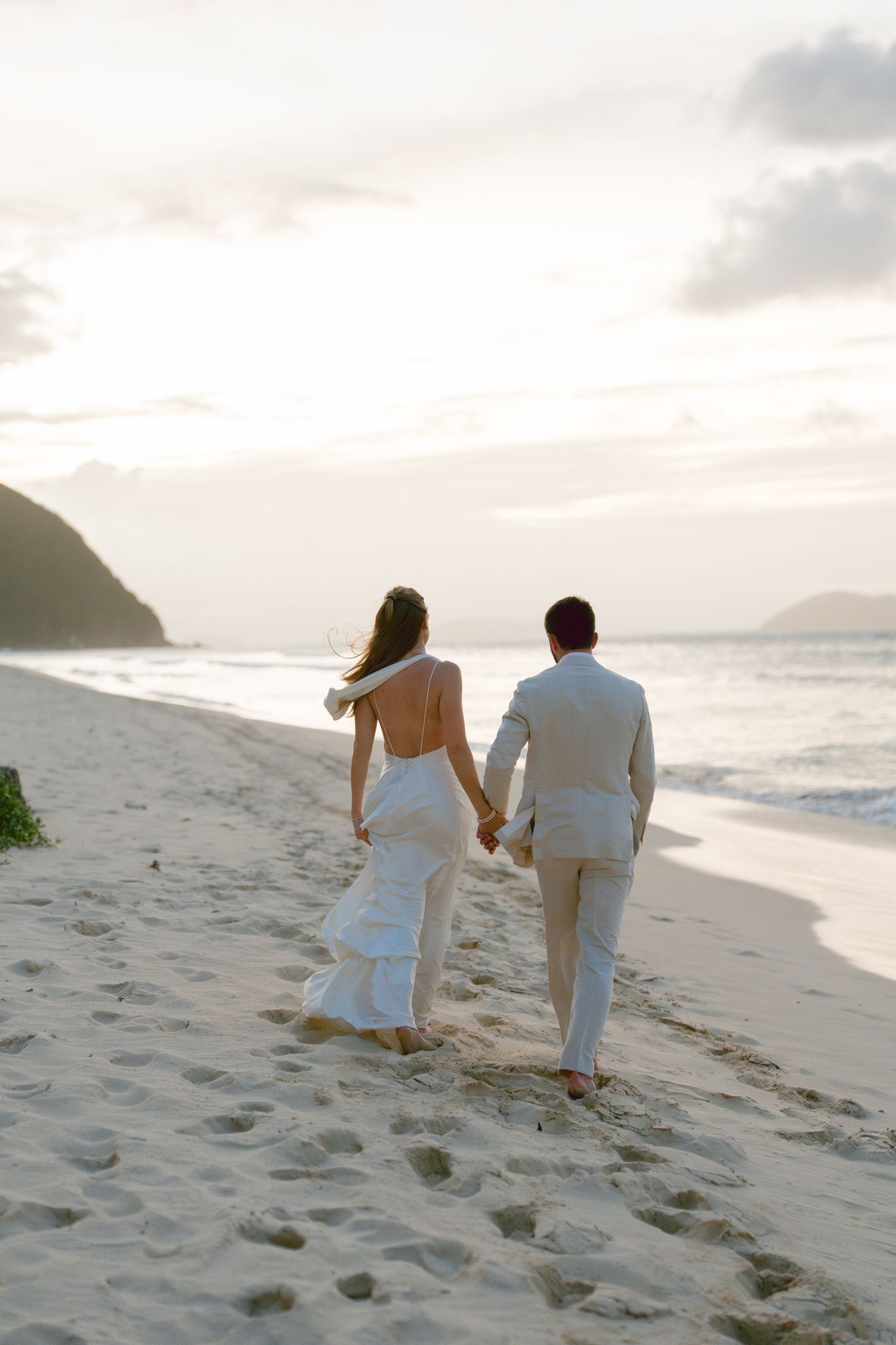 A couple in white attire walking together on a sandy beach at sunset with ocean waves in the background.