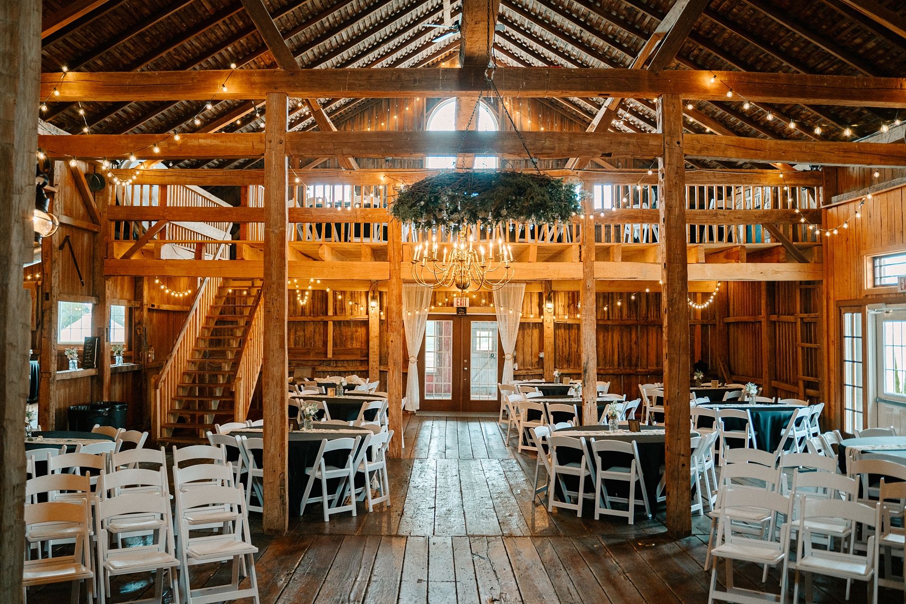Rustic barn wedding reception interior with string lights, chandeliers, and white chairs.