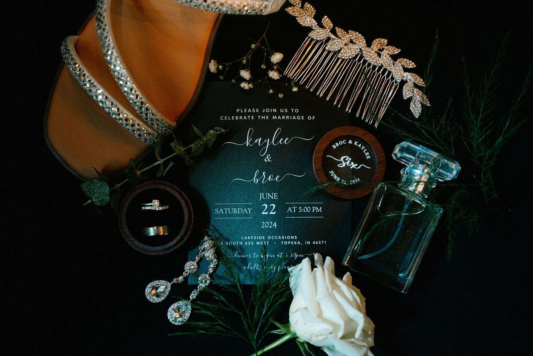Wedding flat lay featuring acrylic invitation, jewelry, perfume, and floral accents.
