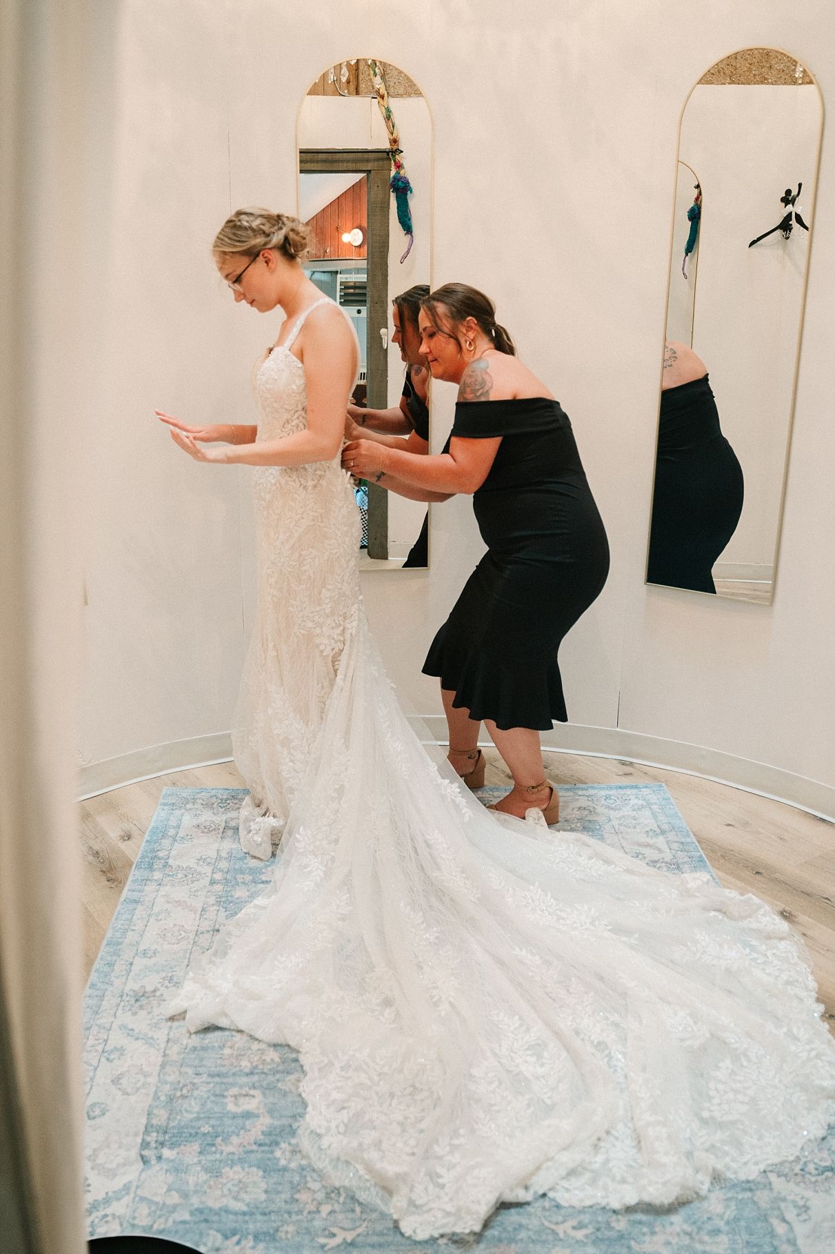 Mother helping bride button up her lace mermaid wedding dress in bridal suite.