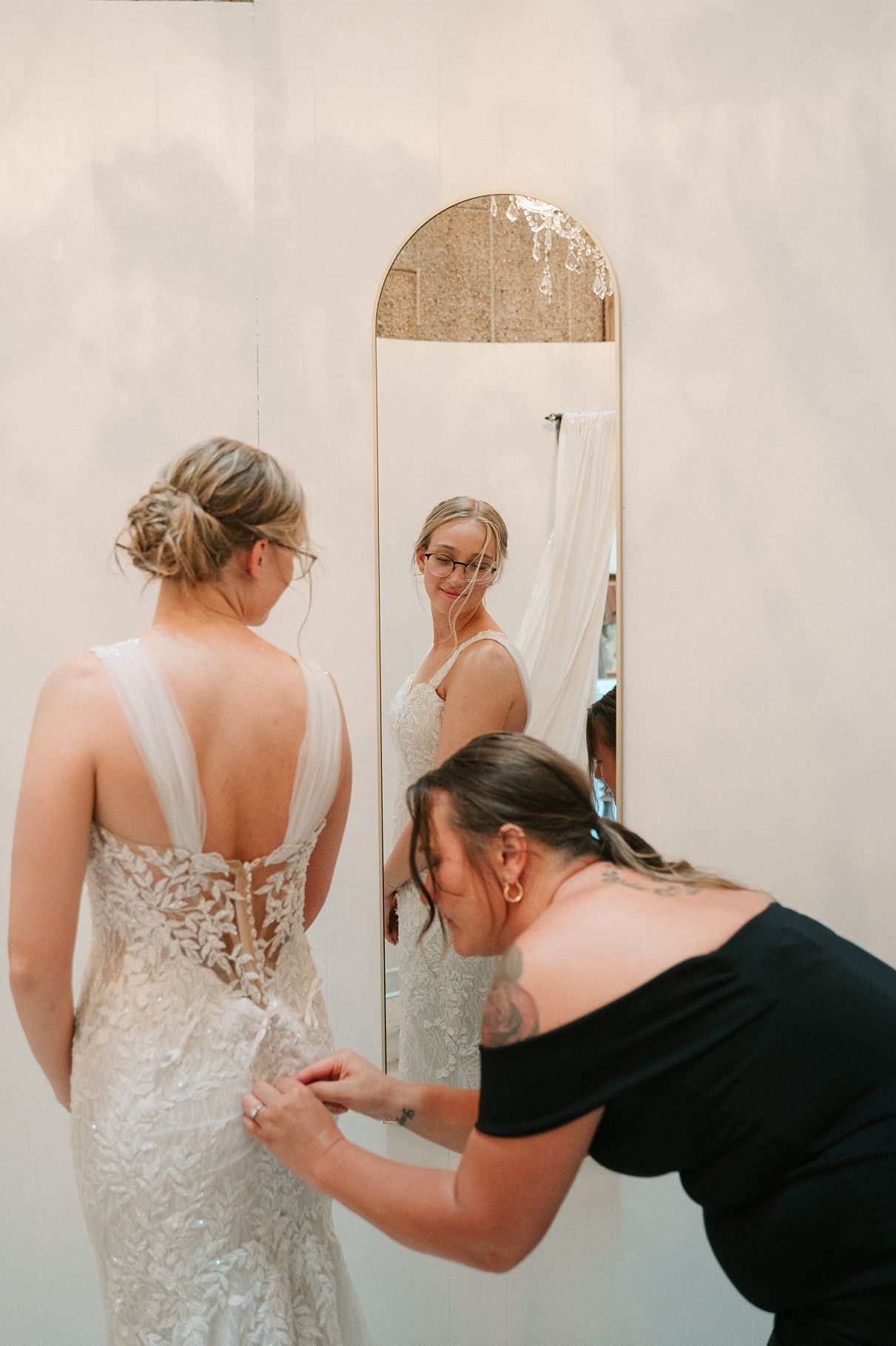 Bridal assistant fastening the back of a lace wedding dress while bride views reflection in gold arch mirror.