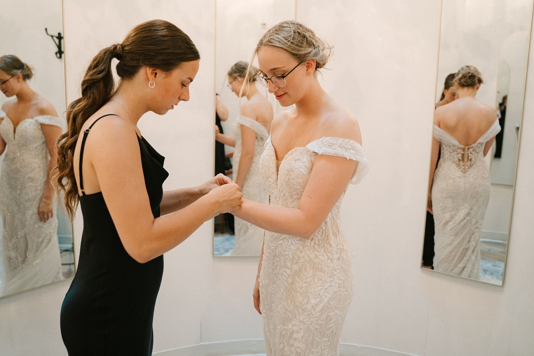 Wedding assistant helping bride in off-shoulder lace gown with finishing touches in a bridal boutique mirror room.
