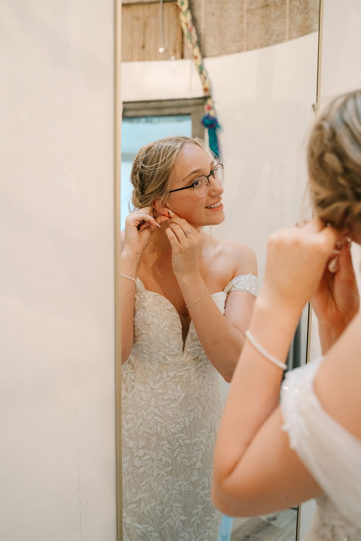 Smiling bride wearing glasses puts on earrings while admiring her lace off-shoulder wedding dress in mirror.